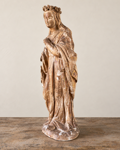 An expressive carved wooden French Antique Madonna, crowned with roses and standing with her gaze gently lowered and one hand resting across her chest. Deeply folded robes and a sweeping mantle give the figure a graceful, sculptural presence, with carved detail continuing around the back.

