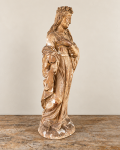 An expressive carved wooden French Antique Madonna, crowned with roses and standing with her gaze gently lowered and one hand resting across her chest. Deeply folded robes and a sweeping mantle give the figure a graceful, sculptural presence, with carved detail continuing around the back.