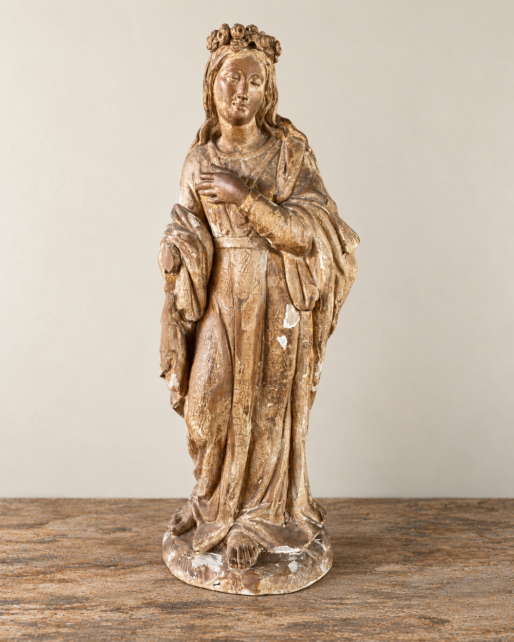 An expressive carved wooden French Antique Madonna, with a crown of roses and standing with her gaze gently lowered and one hand resting across her chest. Deeply folded robes and a sweeping mantle give the figure a graceful, sculptural presence, with carved detail continuing around the back.