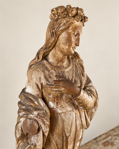 An expressive carved wooden French Antique Madonna, crowned with roses and standing with her gaze gently lowered and one hand resting across her chest. Deeply folded robes and a sweeping mantle give the figure a graceful, sculptural presence, with carved detail continuing around the back.
