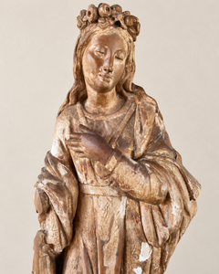 An expressive carved wooden French Antique Madonna, crowned with roses and standing with her gaze gently lowered and one hand resting across her chest. Deeply folded robes and a sweeping mantle give the figure a graceful, sculptural presence, with carved detail continuing around the back.
