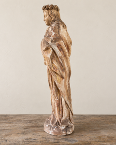 Side view of a French Antique Madonna, crowned with roses and standing with her gaze gently lowered and one hand resting across her chest. Deeply folded robes and a sweeping mantle give the figure a graceful, sculptural presence, with carved detail continuing around the back.