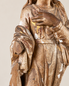 An expressive carved wooden French Antique Madonna, crowned with roses and standing with her gaze gently lowered and one hand resting across her chest. Deeply folded robes and a sweeping mantle give the figure a graceful, sculptural presence, with carved detail continuing around the back.
