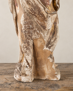An expressive carved wooden French Antique Madonna, crowned with roses and standing with her gaze gently lowered and one hand resting across her chest. Deeply folded robes and a sweeping mantle give the figure a graceful, sculptural presence, with carved detail continuing around the back.

