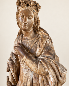 Detail of an expressive carved wooden French Antique Madonna, crowned with roses and standing with her gaze gently lowered and one hand resting across her chest. Deeply folded robes and a sweeping mantle give the figure a graceful, sculptural presence, with carved detail continuing around the back.
